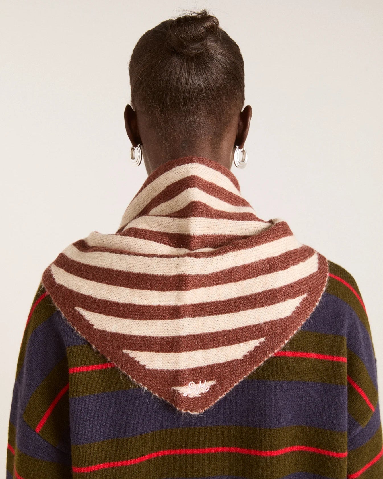 A person shown from the back wearing a striped knit cowl or scarf draped over the shoulders, paired with a dark striped sweater. The cowl features alternating brown and cream stripes, and the sweater has navy, olive green, and red stripes.