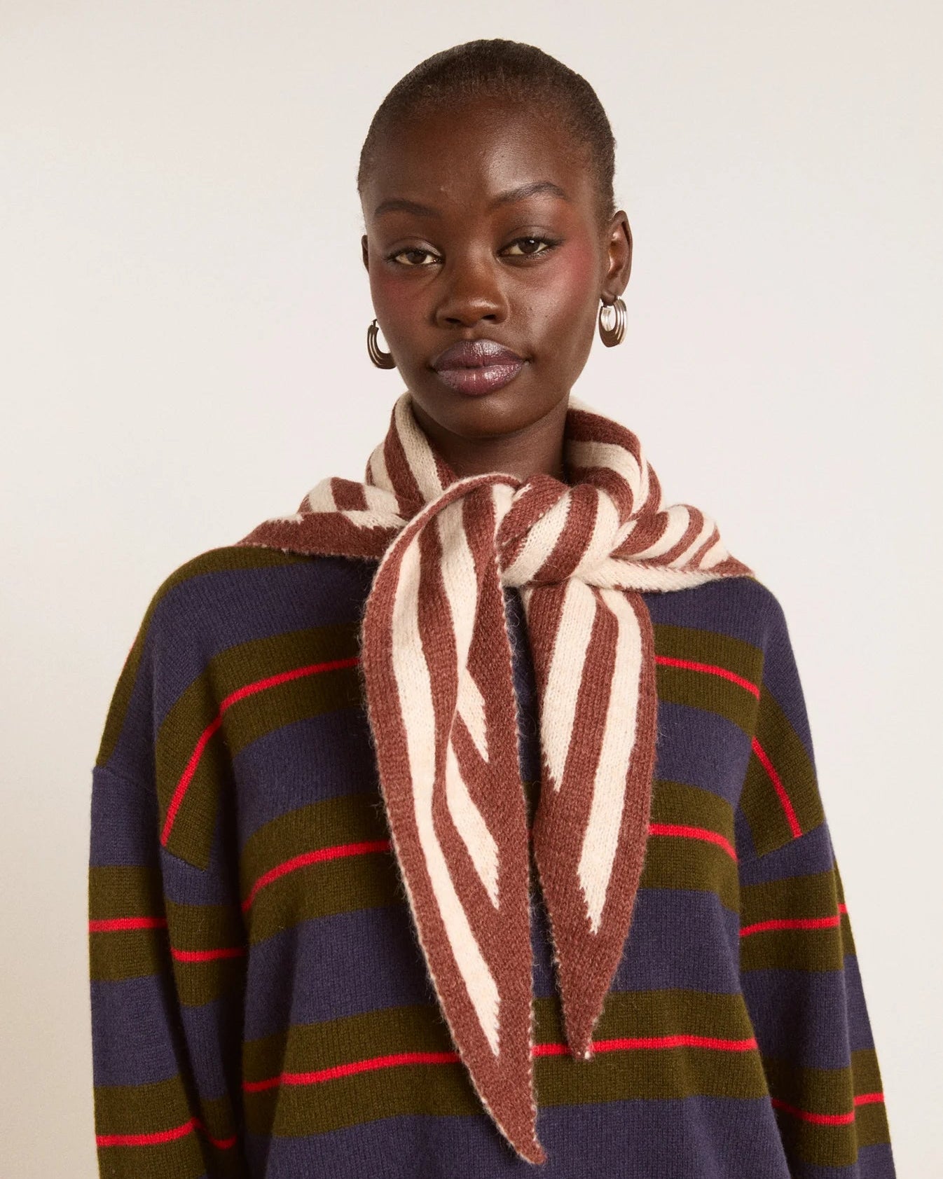 A model wearing a brown and cream striped knit scarf tied around the neck over a navy, olive green, and red striped sweater, against a plain off-white background.