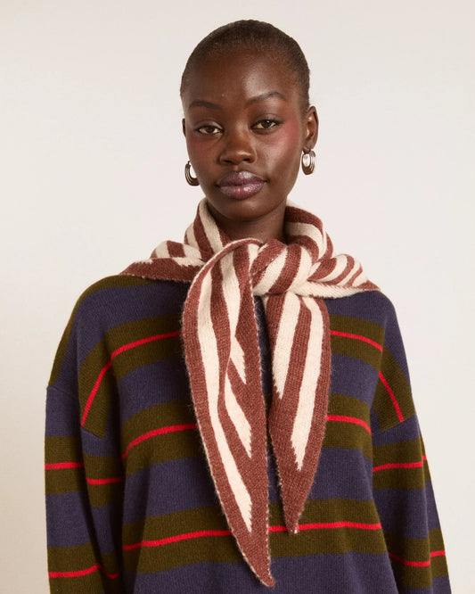 A model wearing a brown and cream striped knit scarf tied around the neck over a navy, olive green, and red striped sweater, against a plain off-white background.